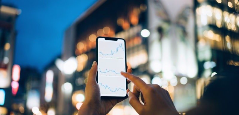 Businesswoman reading financial trading data on smartphone in downtown city street against illuminated urban skyscrapers